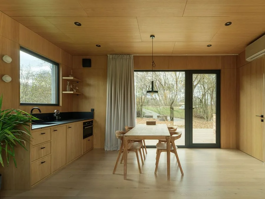 Timber-clad kitchen of a Dwellii cabin: oak fronts, black worktop, dining table, glass door to the deck