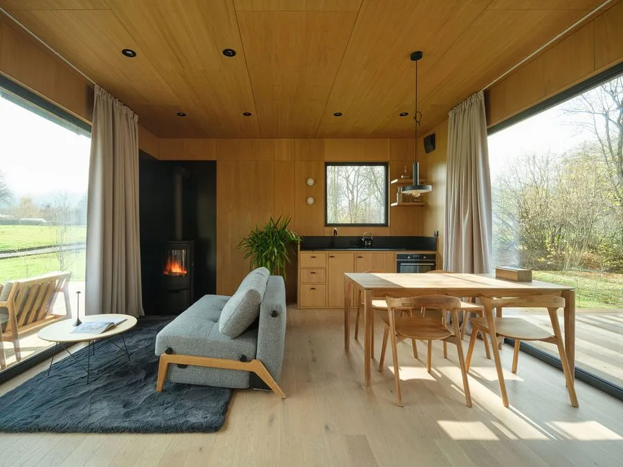 Timber-clad living space of a Dwellii cabin: wood-burning stove, grey sofa, oak dining set, two full-height windows to the meadow