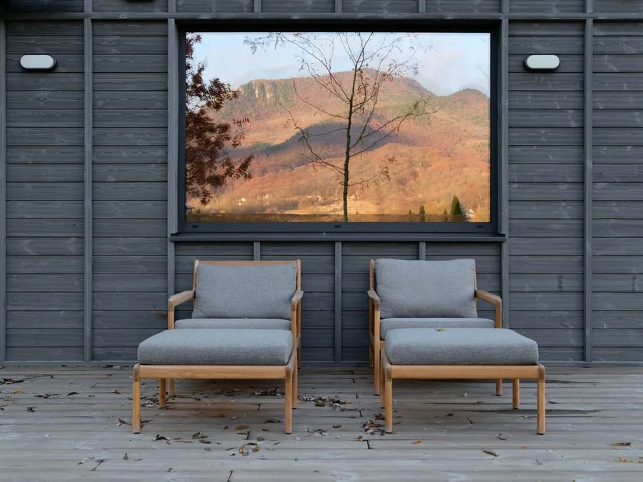 Two upholstered armchairs on the timber deck in front of the grey façade, the mountain ridge reflected in the panoramic window