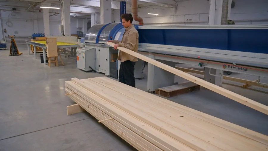 Mitarbeiter gleicht Bauplan, Maßband und Holzbauteil vor dem Zuschnitt ab