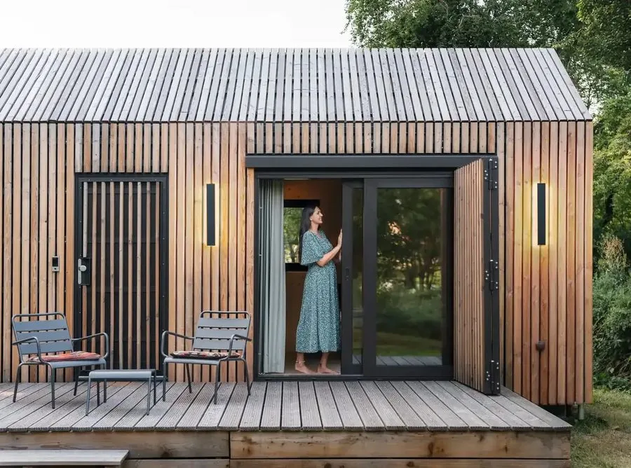 Façade of the Tinca Cabin in detail: larch slats, open sliding door with black frame, terrace chairs, a resident on the deck