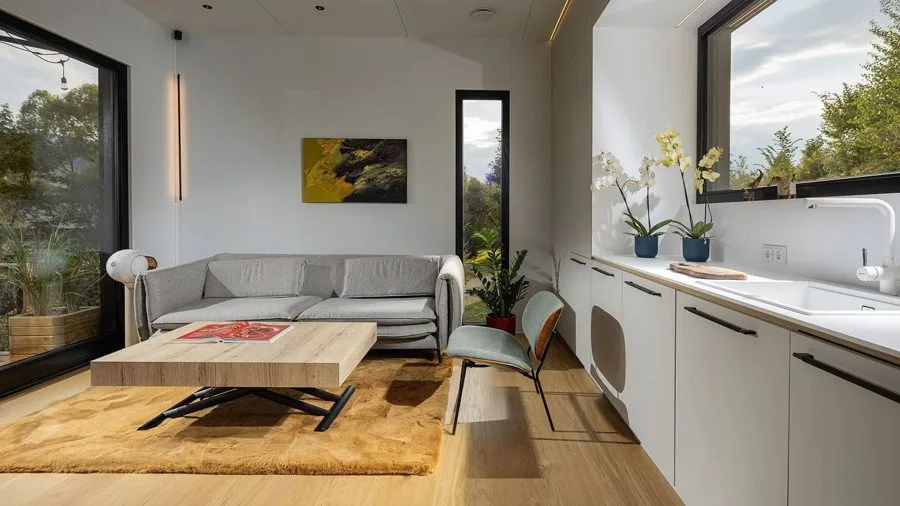Living area of the TR01 with kitchen: grey sofa, coffee table, white fronts, windows on both sides