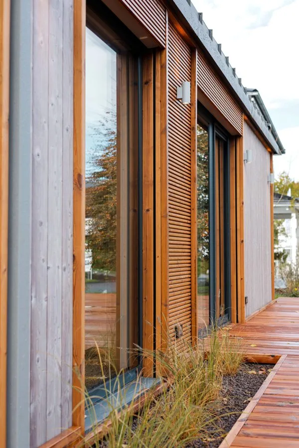 Façade detail of the TR3M: vertical grey larch and warm timber slats, reflections in the glazing, grasses in front