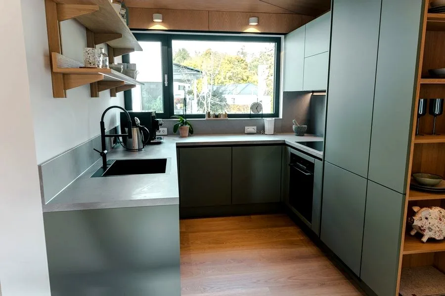 Kitchen in the TR3M showhouse in sage green: window above the sink, built-in oven, open oak shelves, stone worktop